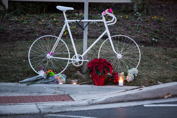 Photo of Tom Palermo memorial; a bike painted white (colloquially known as a 'ghost bike'), and locked to a street post, surrounded by candles, flowers, and other items of remembrance