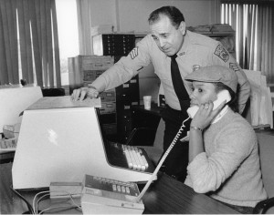 Old photo of a police officer leaning over a woman working on a primitive computer. The woman is taking a phone call.